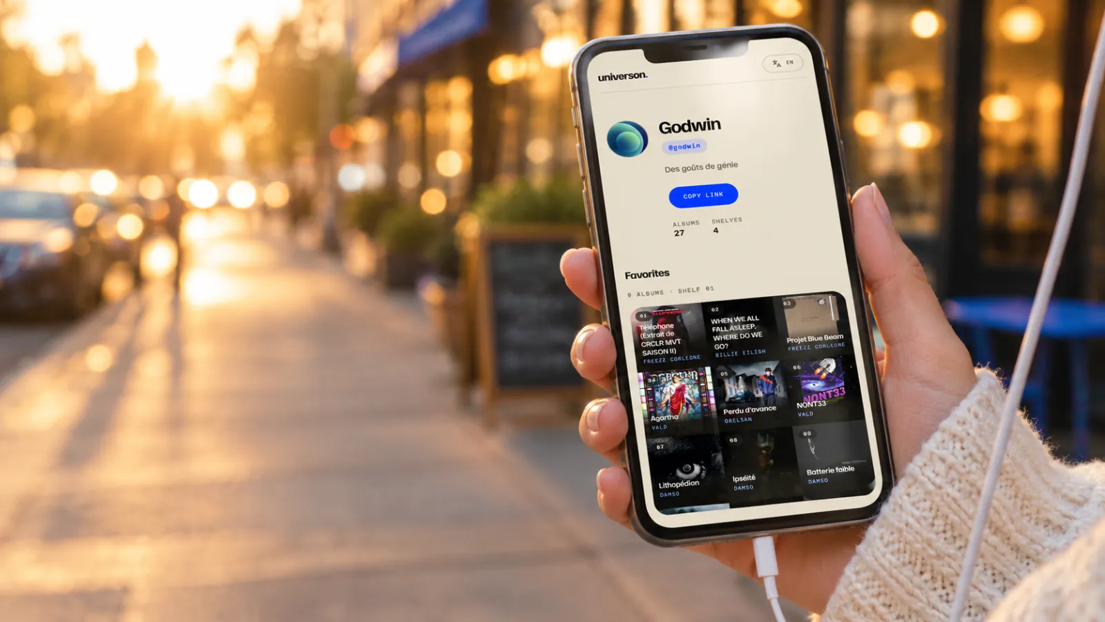 Un téléphone affiche le profil Universon de @godwin dans une rue au coucher du soleil